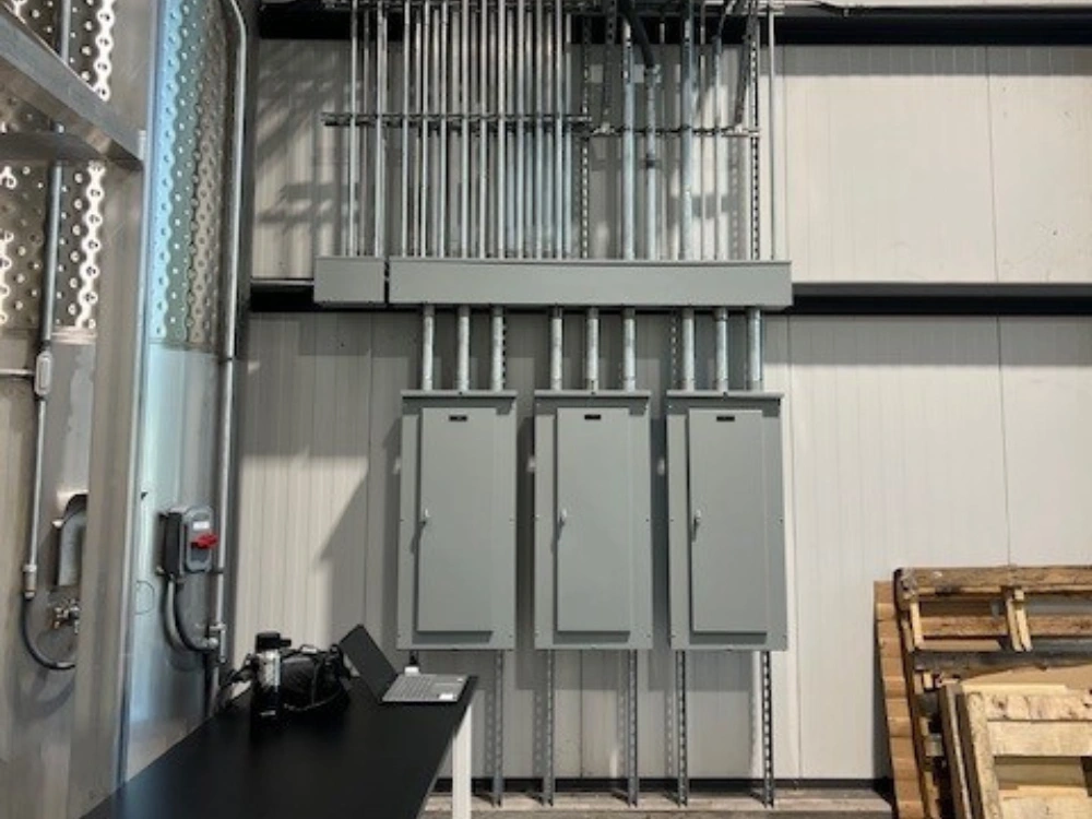 Industrial electrical installation featuring three vertical wall-mounted gray breaker panels with extensive metal conduit piping running up to the high ceiling.