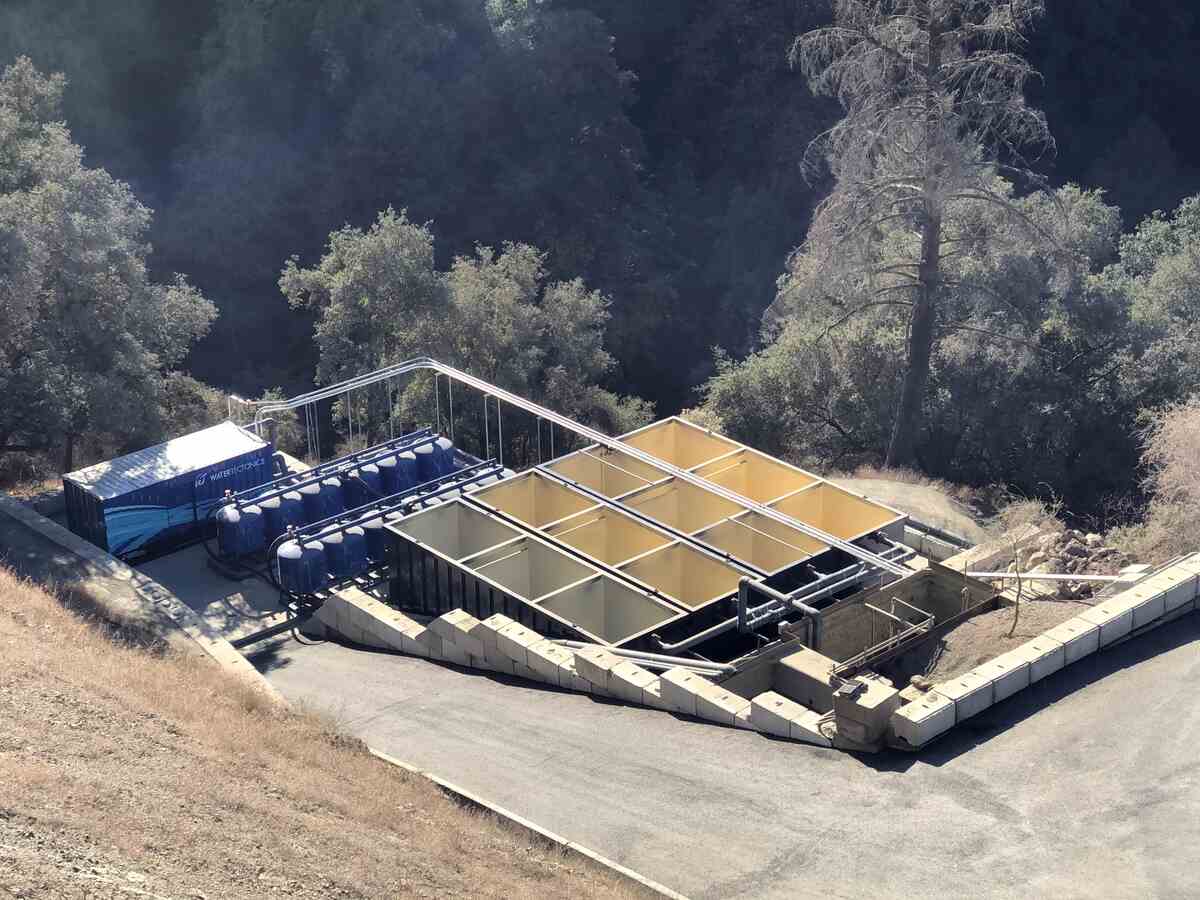 Aerial view of an outdoor industrial storm water treatment facility by Knight's Electric, featuring rectangular settling basins, blue filtration tanks, and a WaterTectonics control container surrounded by trees.
